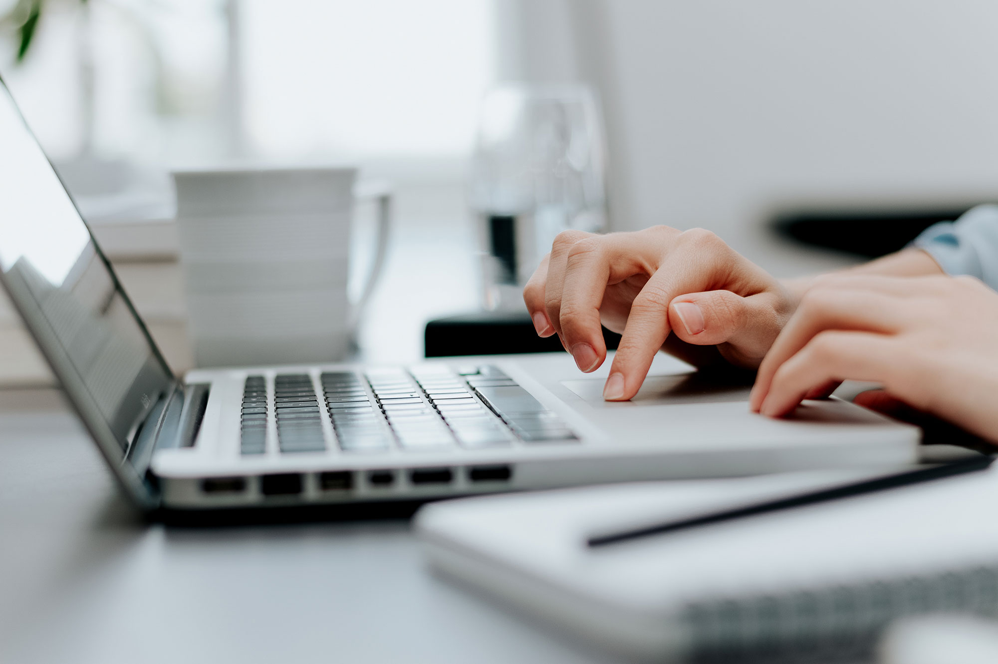 Close-up of hands working on a laptop with a pen and paper in the foreground.